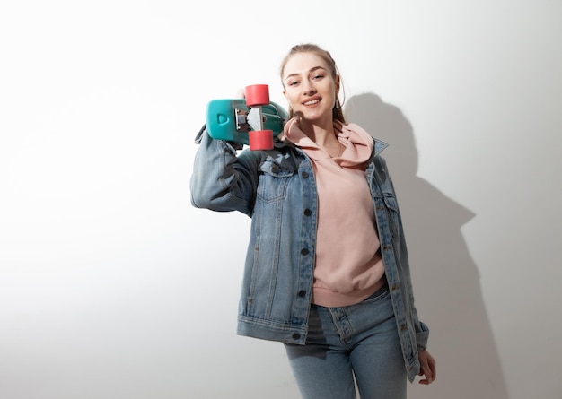 Jonge blanke vrolijke hipster vrouw in denim kleding met penny board witte achtergrond met lange schaduw