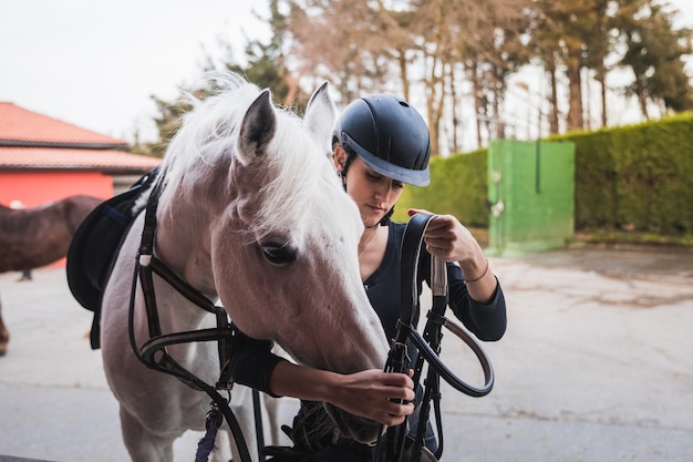 Jonge blanke vrouw bereidt een wit paard voor op een ritje