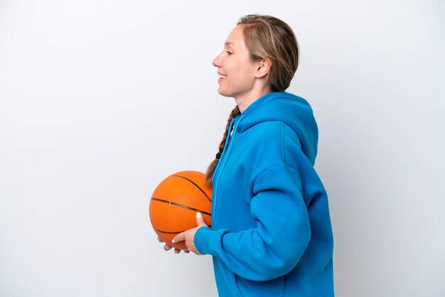 Jonge blanke vrouw die basketbal speelt geïsoleerd op een witte achtergrond lachend in zijpositie