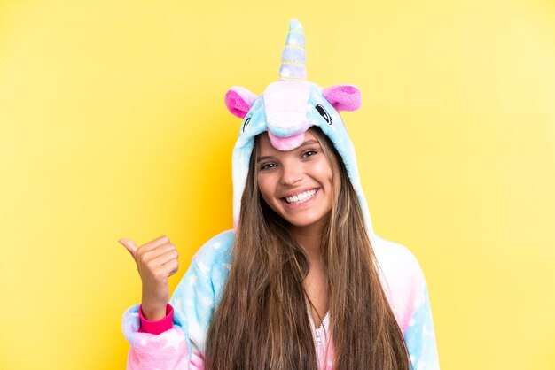 Jonge blanke vrouw die een eenhoornpyjama draagt, geïsoleerd op een gele achtergrond, wijzend naar de zijkant om een product te presenteren