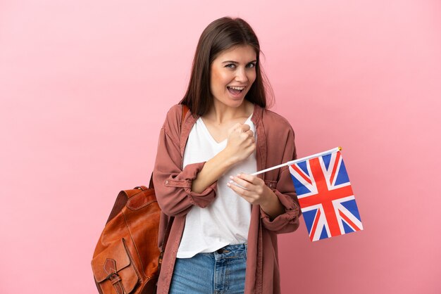 Jonge blanke vrouw die een vlag van het Verenigd Koninkrijk houdt die op roze achtergrond wordt geïsoleerd die een overwinning viert