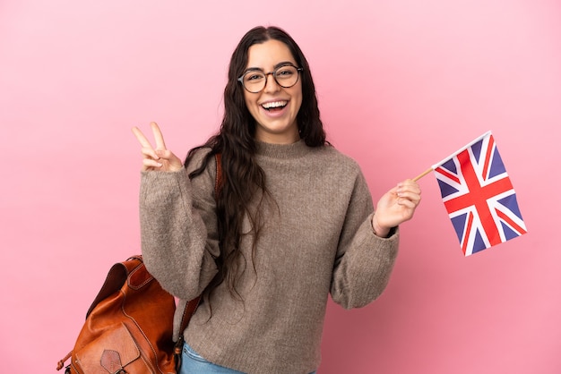 Jonge blanke vrouw die een vlag van het Verenigd Koninkrijk houdt die op roze achtergrond wordt geïsoleerd die en overwinningsteken glimlacht toont