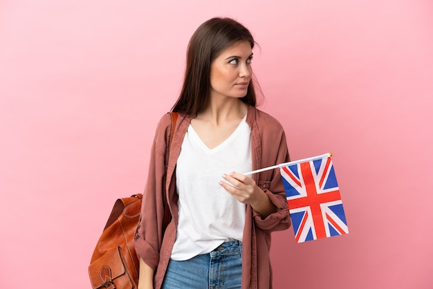 Jonge blanke vrouw die een vlag van het Verenigd Koninkrijk houdt die op roze achtergrond wordt geïsoleerd en naar de zijkant kijkt