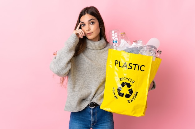 Jonge blanke vrouw die een zak vol plastic flessen houdt om te recyclen geïsoleerd op roze achtergrond, denkend een idee