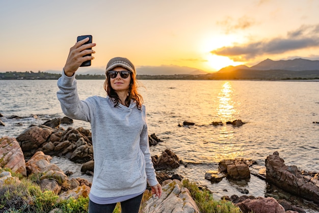 Jonge blanke vrouw die een zelfportret met zonnebril en wollen hoed op ocean sea resort bij zonsondergang of zonsopgang. Vrouwelijke soloreiziger die plezier heeft met het delen van haar foto op sociaal netwerk