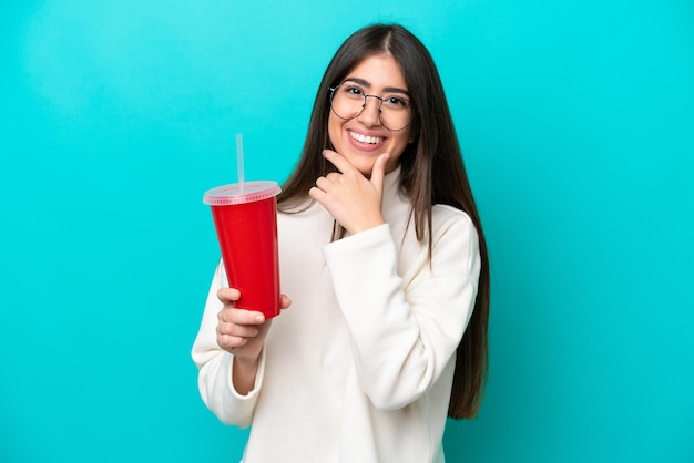 Jonge blanke vrouw die frisdrank drinkt geïsoleerd op blauwe achtergrond gelukkig en glimlachend