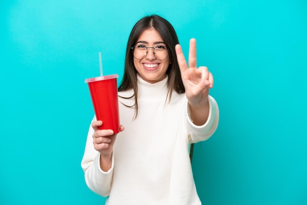 Jonge blanke vrouw die frisdrank drinkt geïsoleerd op blauwe achtergrond glimlachend en overwinningsteken toont