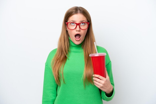 Jonge blanke vrouw die frisdrank drinkt geïsoleerd op een witte achtergrond die omhoog kijkt en met een verbaasde uitdrukking