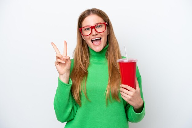 Jonge blanke vrouw die frisdrank drinkt geïsoleerd op een witte achtergrond glimlachend en overwinningsteken toont