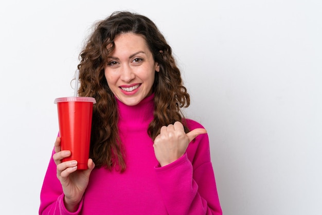 Jonge blanke vrouw die frisdrank drinkt geïsoleerd op een witte achtergrond, wijzend naar de zijkant om een product te presenteren