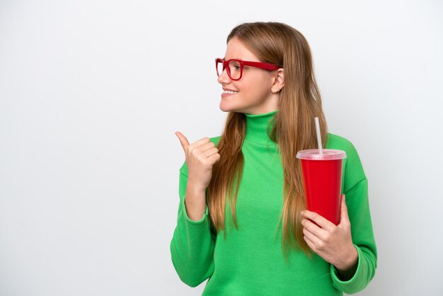 Jonge blanke vrouw die frisdrank drinkt geïsoleerd op een witte achtergrond, wijzend naar de zijkant om een product te presenteren