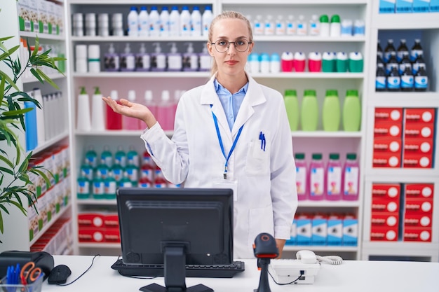 Jonge blanke vrouw die in een apotheek werkt, glimlacht vrolijk, presenteert en wijst met de handpalm naar de camera.