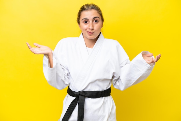 Jonge blanke vrouw die karate doet geïsoleerd op gele achtergrond die twijfels heeft terwijl ze haar handen opsteekt