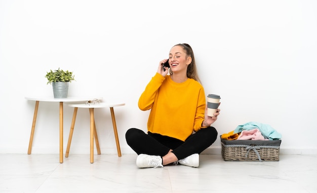 Jonge blanke vrouw die kleding vouwt die op de vloer zit geïsoleerd op een witte achtergrond met koffie om mee te nemen en een mobiel