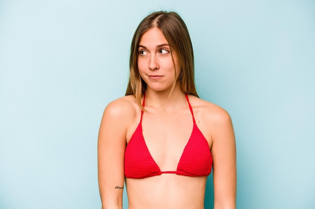 Jonge blanke vrouw die naar het strand gaat geïsoleerd op een blauwe achtergrond verward voelt zich twijfelachtig en onzeker