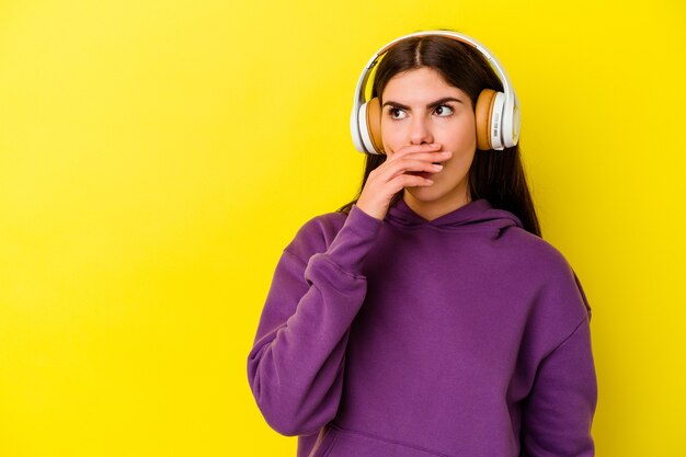 Jonge blanke vrouw die naar muziek luistert met een koptelefoon geïsoleerd op een roze achtergrond, bedachtzaam op zoek naar een kopieerruimte die de mond bedekt met de hand.