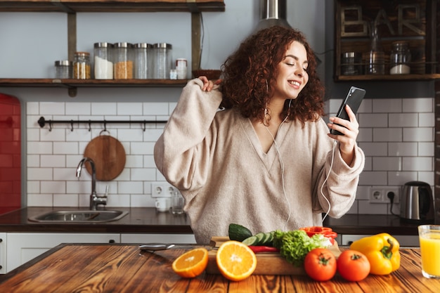 jonge blanke vrouw die naar muziek luistert op mobiele telefoon tijdens het koken van verse groentensalade in het keukeninterieur thuis