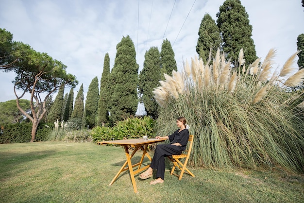 Jonge blanke vrouw die op laptopcomputer werkt en thee drinkt terwijl ze aan een houten tafel zit op een prachtig groen mediterraan gazon. Concept van weekend, rust en werken op afstand