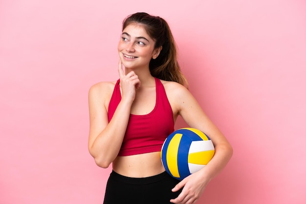 Jonge blanke vrouw die volleybal speelt geïsoleerd op roze achtergrond en een idee denkt terwijl ze omhoog kijkt
