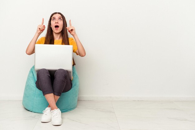 Jonge blanke vrouw die werkt met laptop zittend op een trekje geïsoleerd op een witte achtergrond wijzend ondersteboven met geopende mond.