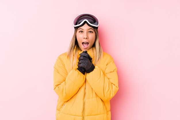 Jonge blanke vrouw draagt een ski-kleding in een roze muur bidden voor geluk, verbaasd en mond openen