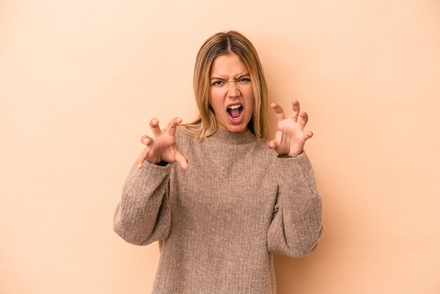 Jonge blanke vrouw geïsoleerd op beige achtergrond boos schreeuwen met gespannen handen.