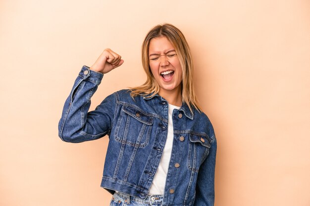 Jonge blanke vrouw geïsoleerd op beige achtergrond die een overwinning, passie en enthousiasme, gelukkige uitdrukking viert.