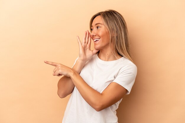 Jonge blanke vrouw geïsoleerd op beige achtergrond die een roddel zegt, wijzend naar de zijkant die iets meldt.