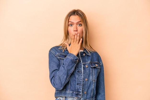 Jonge blanke vrouw geïsoleerd op beige achtergrond geschokt, mond bedekken met handen, angstig om iets nieuws te ontdekken.