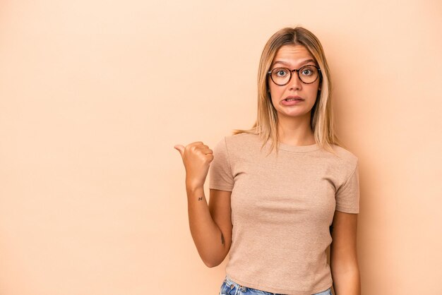 Jonge blanke vrouw geïsoleerd op beige achtergrond geschokt wijzend met wijsvingers naar een kopieerruimte.