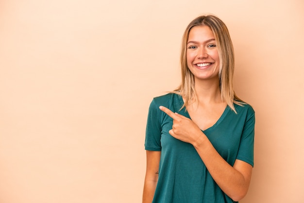 Jonge blanke vrouw geïsoleerd op beige achtergrond glimlachend en opzij wijzend, iets tonend op lege ruimte.