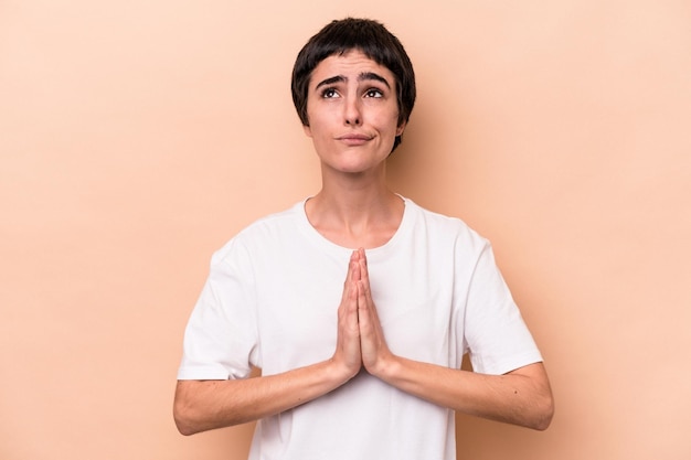 Jonge blanke vrouw geïsoleerd op beige achtergrond hand in hand bidden in de buurt van mond, voelt zich zelfverzekerd.