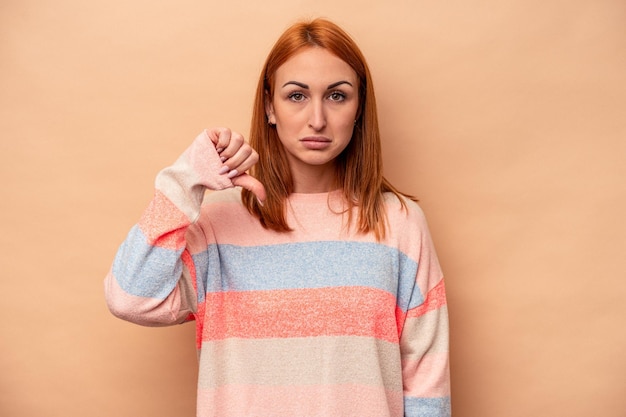 Jonge blanke vrouw geïsoleerd op beige achtergrond met een afkeer gebaar duimen omlaag meningsverschil concept