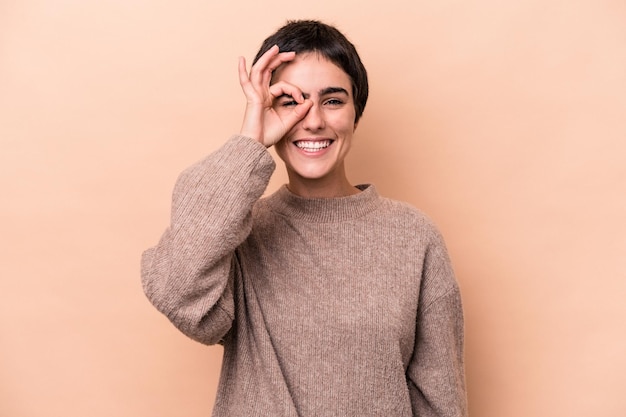 Jonge blanke vrouw geïsoleerd op beige achtergrond opgewonden houden ok gebaar op oog.