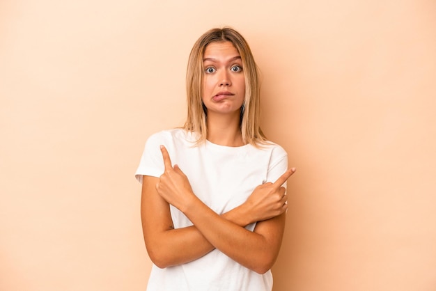 Jonge blanke vrouw geïsoleerd op beige achtergrond wijst zijwaarts, probeert te kiezen tussen twee opties.