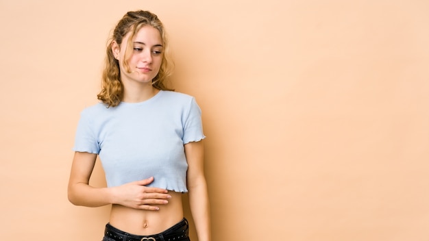 Jonge blanke vrouw geïsoleerd op beige muur raakt buik, glimlacht zachtjes, eten en tevredenheid concept.