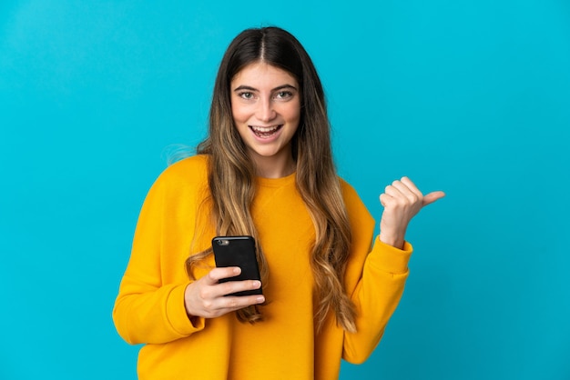 Jonge blanke vrouw geïsoleerd op blauw met behulp van mobiele telefoon en wijst naar de zijkant