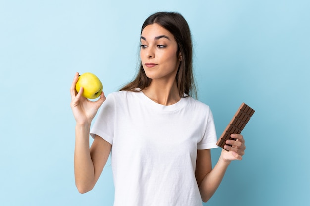 Jonge blanke vrouw geïsoleerd op blauw met twijfels tijdens het nemen van een chocoladetablet in de ene hand en een appel in de andere