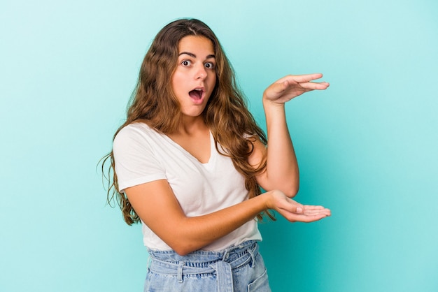 Jonge blanke vrouw geïsoleerd op blauwe achtergrond geschokt en verbaasd met een kopie ruimte tussen handen.