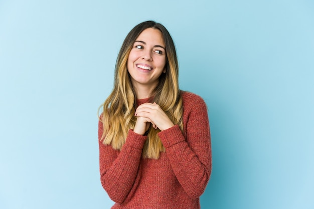 Jonge blanke vrouw geïsoleerd op blauwe achtergrond houdt de handen onder de kin, kijkt gelukkig opzij.