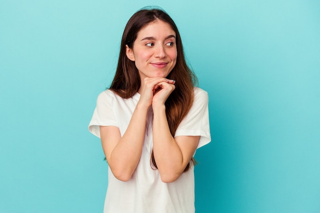 Jonge blanke vrouw geïsoleerd op blauwe achtergrond houdt handen onder de kin, kijkt vrolijk opzij.