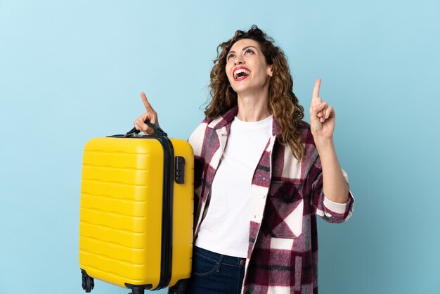 Jonge blanke vrouw geïsoleerd op blauwe achtergrond in vakantie met reiskoffer en omhoog