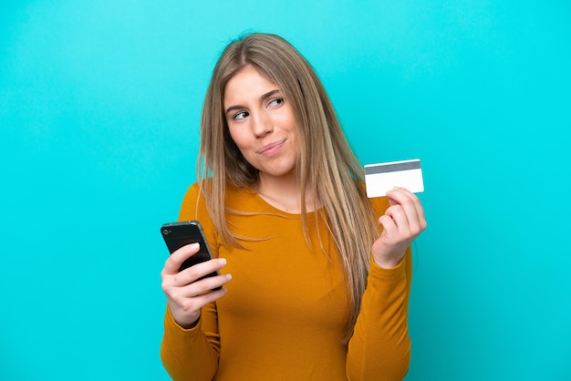 Jonge blanke vrouw geïsoleerd op blauwe achtergrond kopen met de mobiel met een creditcard terwijl ze denken while