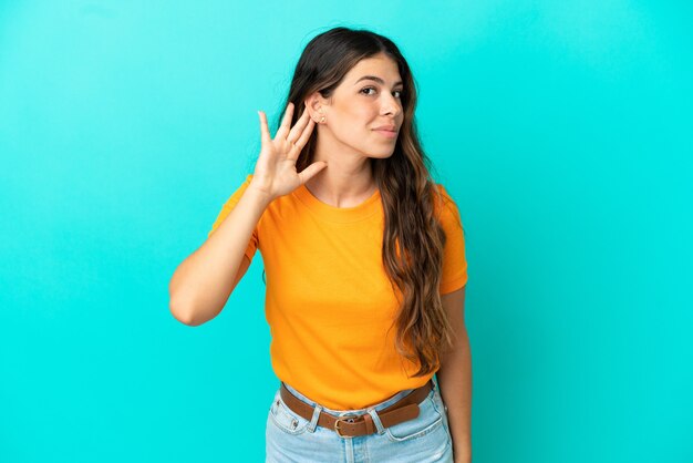 Jonge blanke vrouw geïsoleerd op blauwe achtergrond luisteren naar iets door hand op het oor te leggen