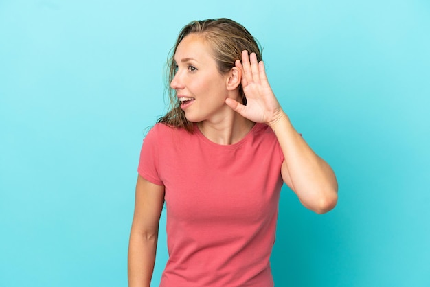 Jonge blanke vrouw geïsoleerd op blauwe achtergrond luisteren naar iets door hand op het oor te leggen