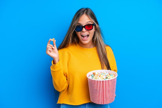 Jonge blanke vrouw geïsoleerd op blauwe achtergrond met 3D-bril en met een grote emmer popcorns