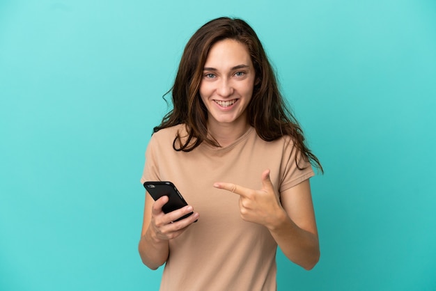 Jonge blanke vrouw geïsoleerd op blauwe achtergrond met behulp van mobiele telefoon en erop te wijzen