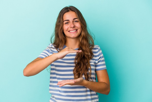 Jonge blanke vrouw geïsoleerd op blauwe achtergrond met iets met beide handen, productpresentatie.