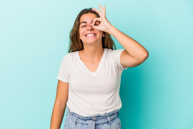 Jonge blanke vrouw geïsoleerd op blauwe achtergrond opgewonden houden ok gebaar op oog.