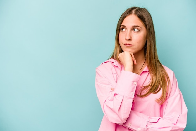 Jonge blanke vrouw geïsoleerd op blauwe achtergrond zijwaarts kijkend met twijfelachtige en sceptische uitdrukking